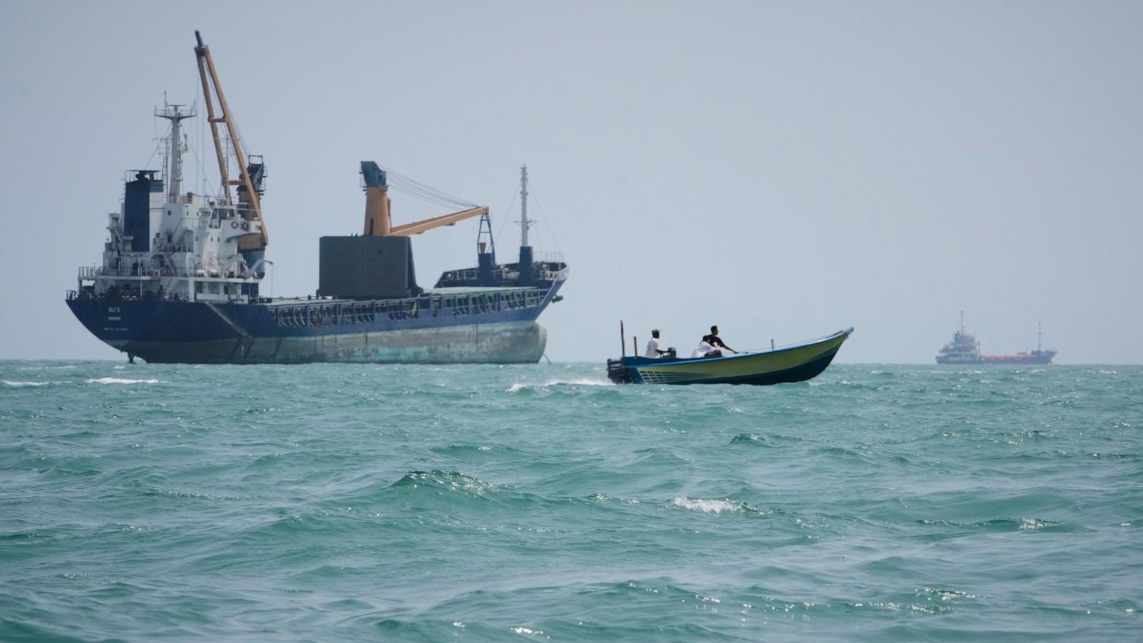A large cargo ship and a small motorboat carrying several people travel across choppy turquoise water.