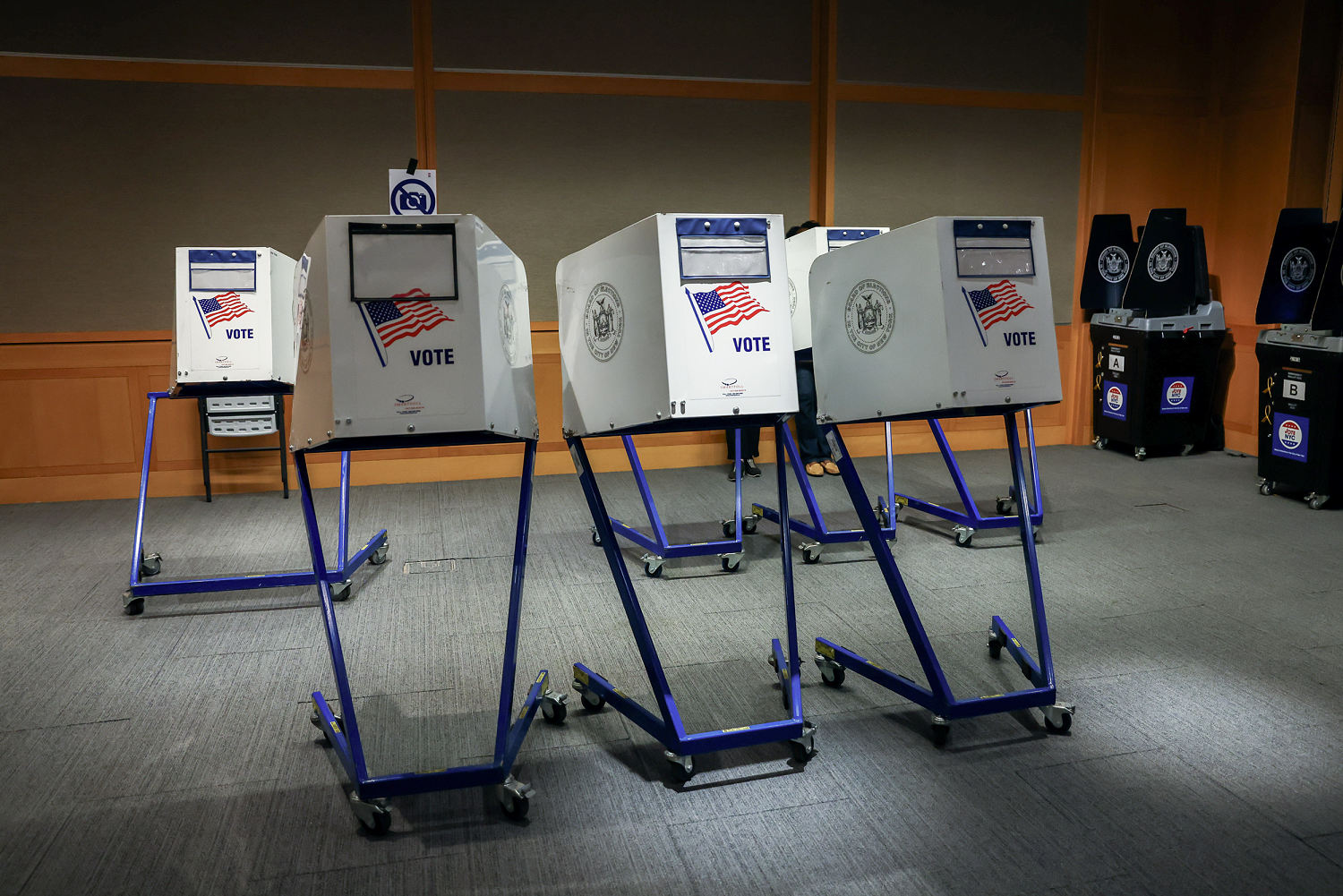 Several wheeled voting booths and electronic voting machines stand inside a wood-paneled polling place.