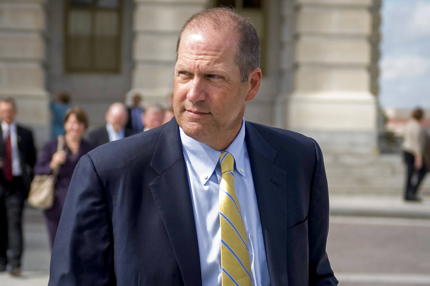 Sununu walks outdoors in a dark suit and striped yellow tie, with several people and a stone building behind him.