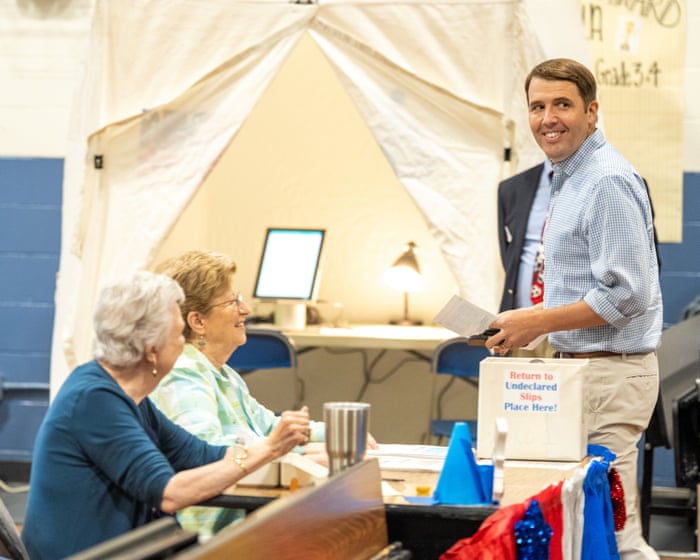 Pappas stands holding papers beside seated election workers at a table decorated with red, white, and blue bunting.