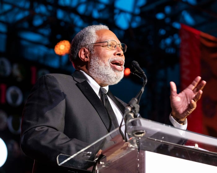 Lonnie Bunch gestures while speaking into a microphone from a clear lectern on a stage.