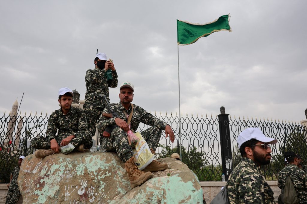 Armed men in camouflage gather beside a metal fence beneath a green flag.