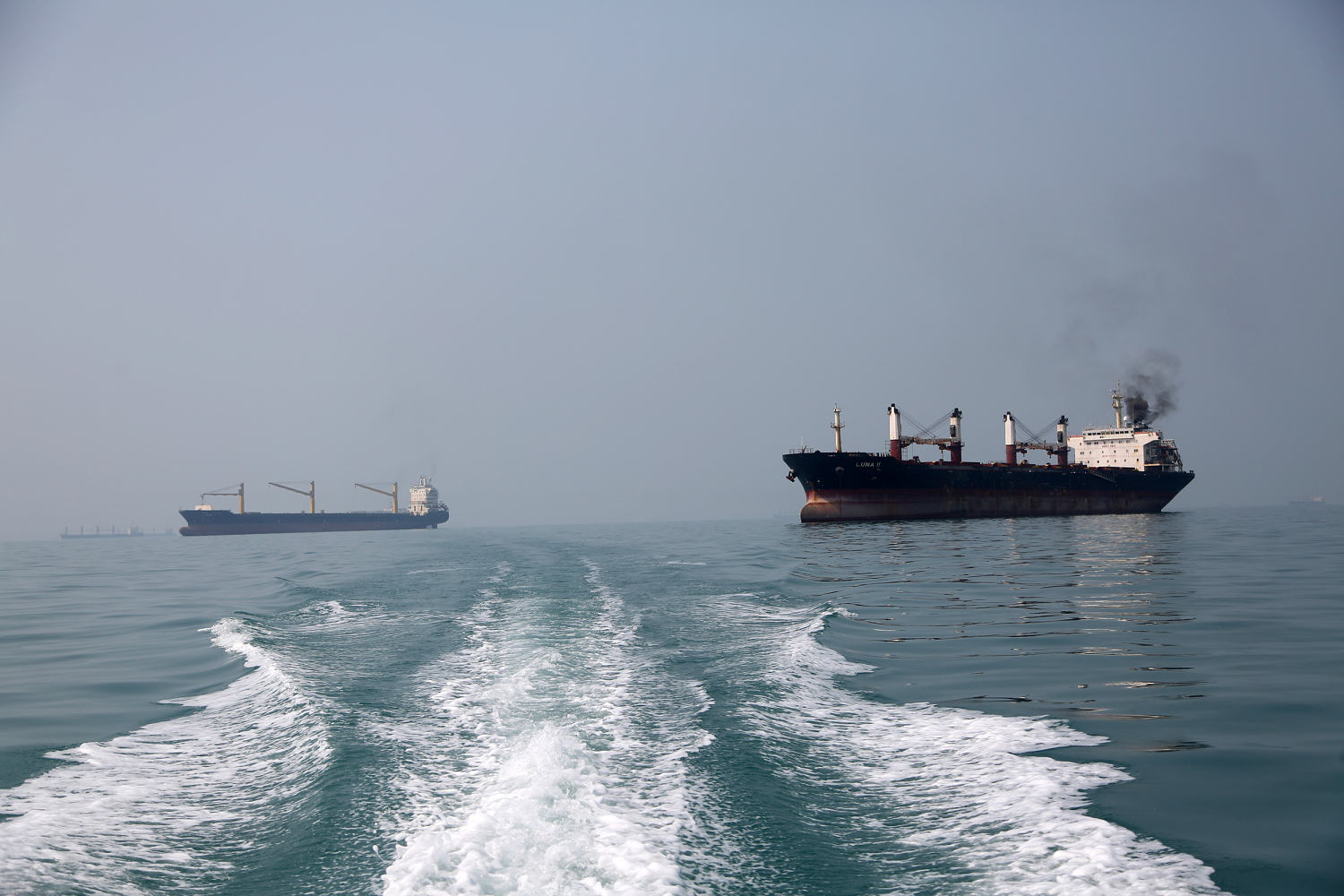 Two cargo ships on open water, seen beyond a boat’s wake.