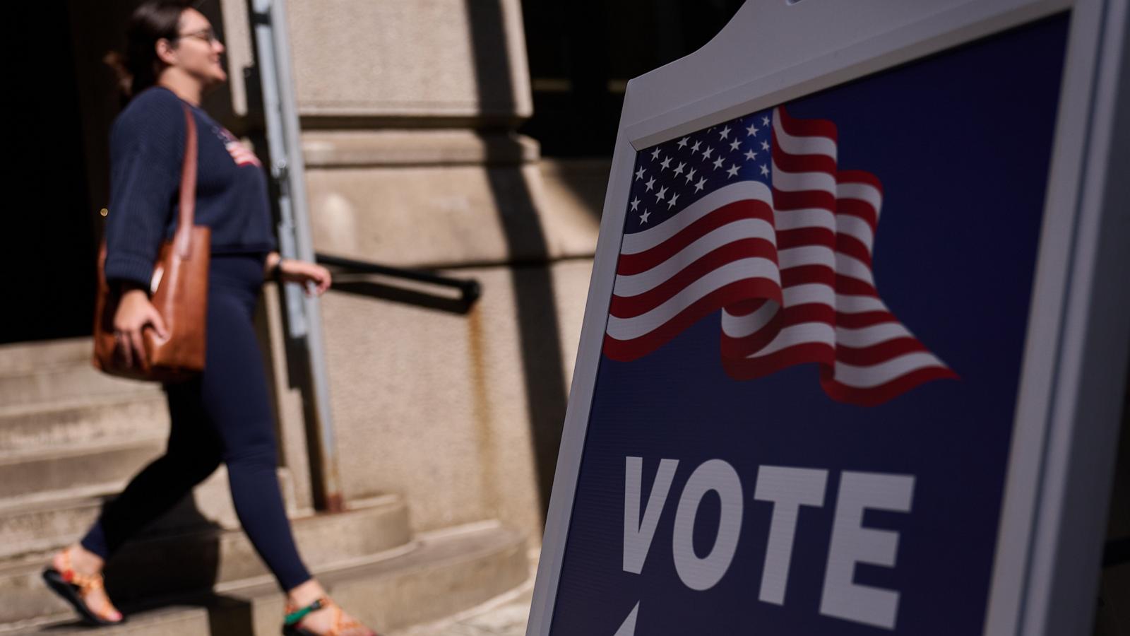A woman walks past a sidewalk VOTE sign bearing an American flag.