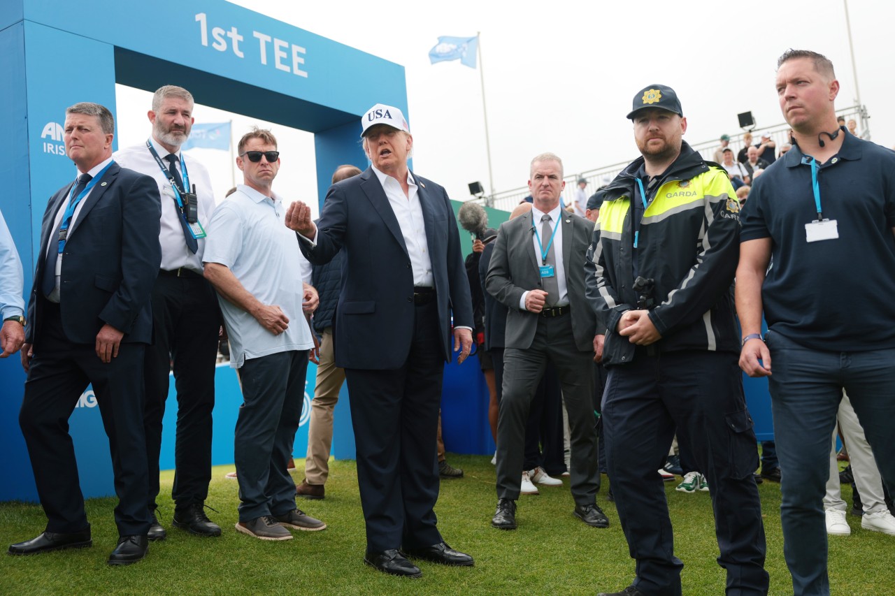 Trump gestures while standing among security personnel and other attendees beside a blue first-tee structure at a golf event.