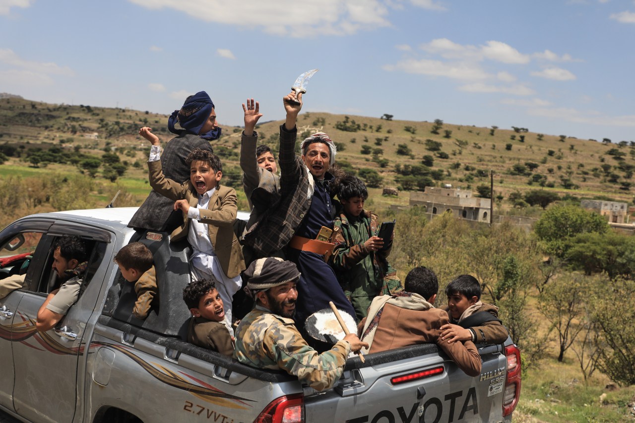 Men and boys crowd into a pickup truck, raising their hands and holding a curved dagger and drum.