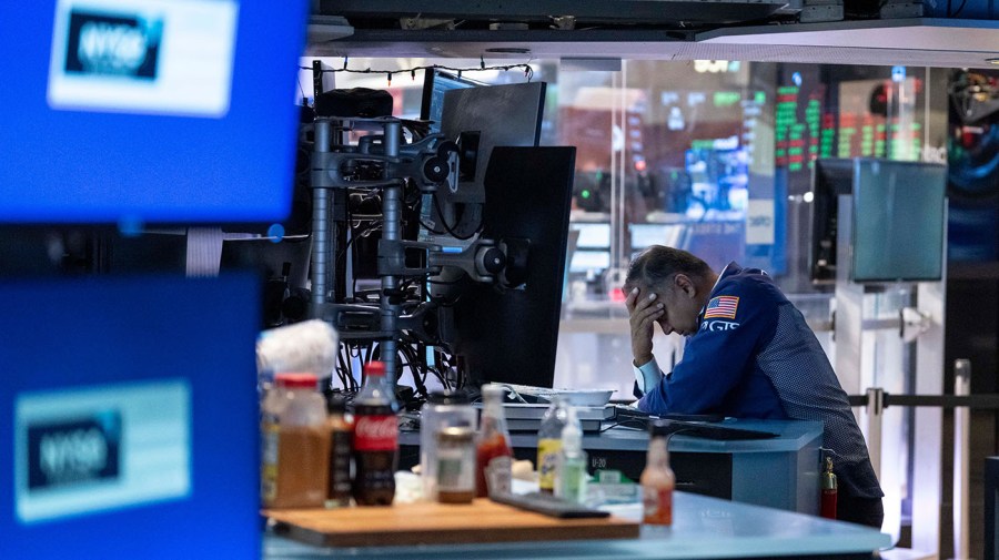 A seated trader covers his face with one hand beside monitors on the floor of a stock exchange.