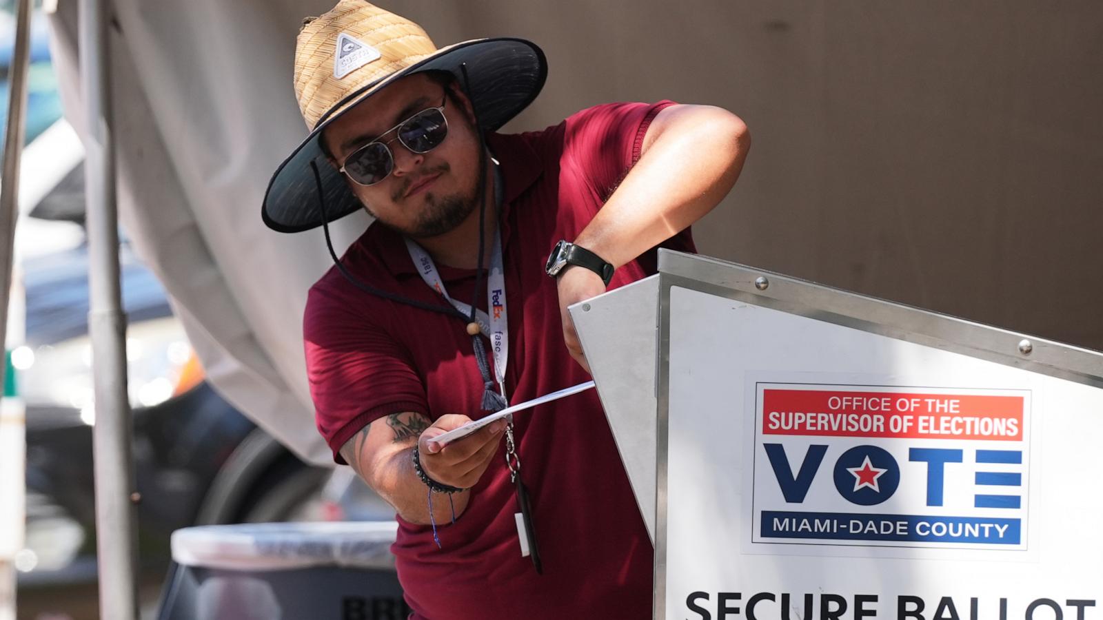 A voter in a straw hat and sunglasses drops a ballot envelope into a Miami-Dade County secure ballot box.