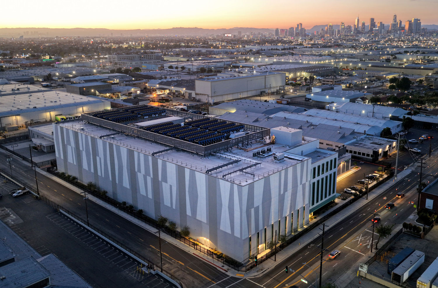 A windowless industrial data-center building with rooftop cooling equipment amid warehouses, a city skyline in the distance.