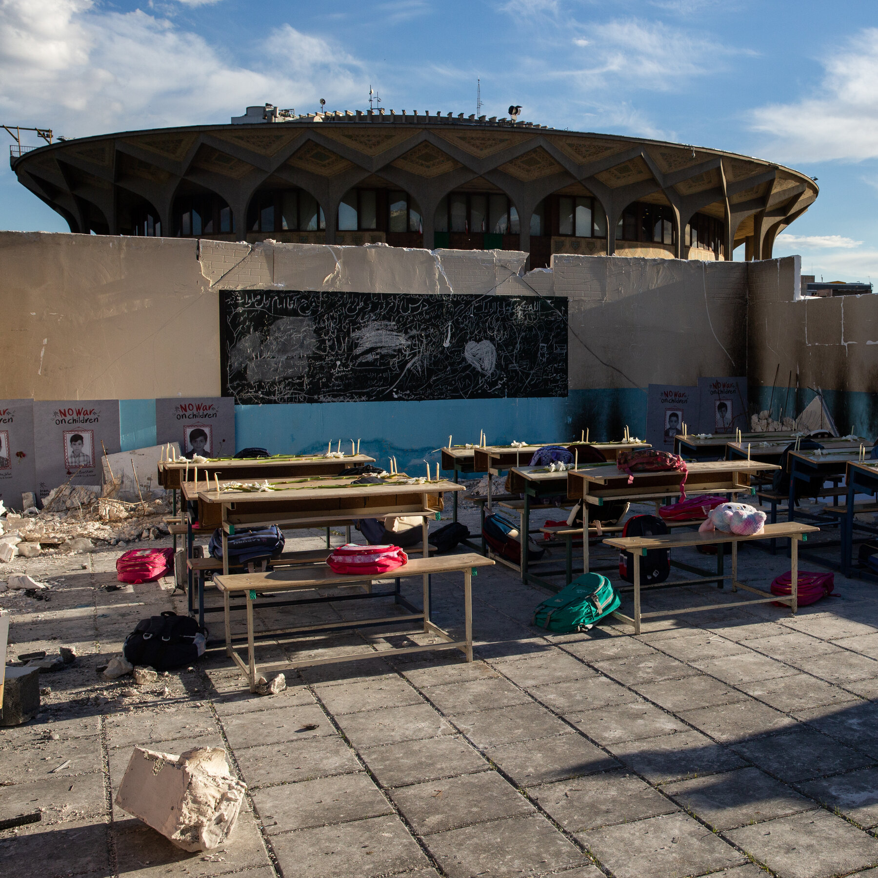 Children's backpacks and shoes rest on classroom desks beneath a chalkboard and a 'no war on children' poster inside the damaged school.