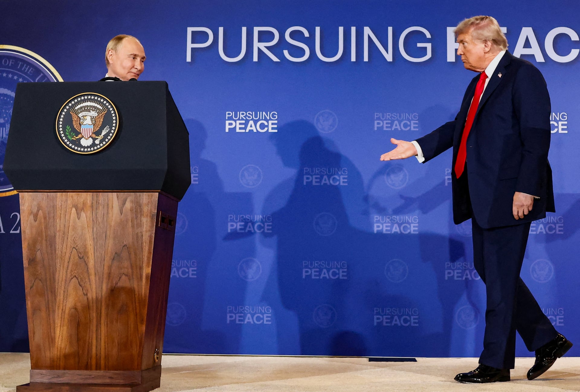 President Trump reaches toward Vladimir Putin on a stage beneath a sign reading 'Pursuing Peace.'
