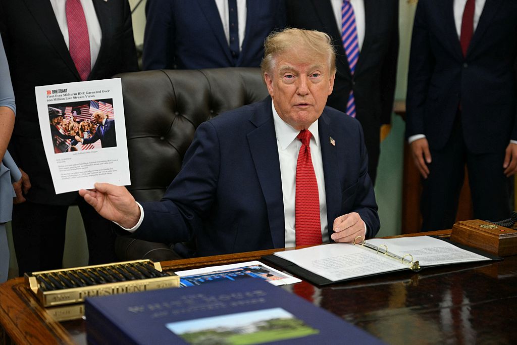 President Trump sits at the Resolute desk in the Oval Office holding up a printed page of favorable coverage.