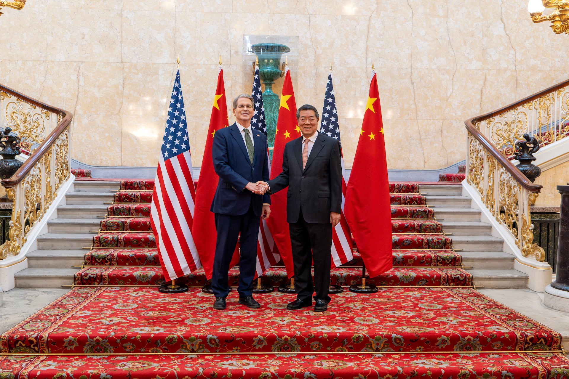 Treasury Secretary Scott Bessent and Vice Premier He Lifeng shake hands on a staircase between U.S. and Chinese flags.