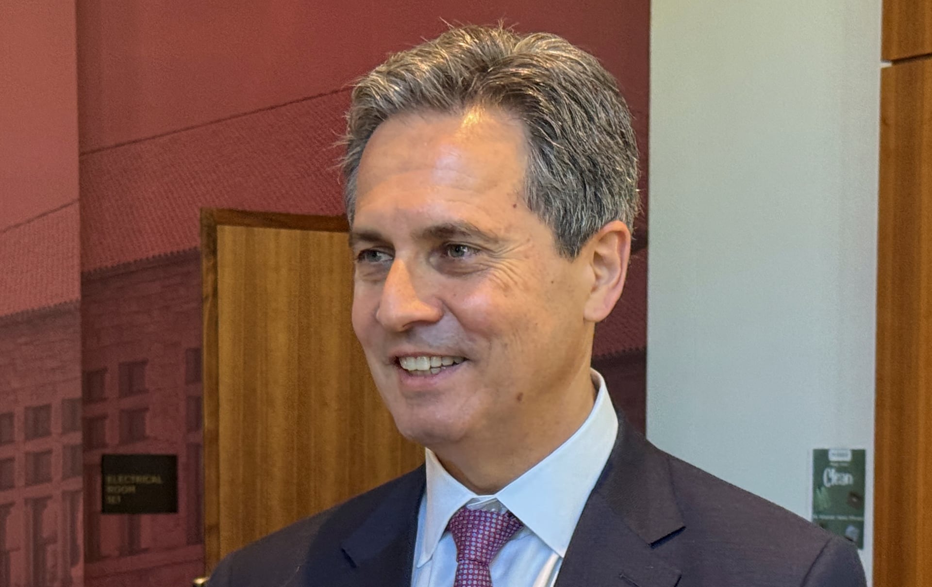 Austan Goolsbee smiles while wearing a dark suit and patterned tie inside a wood-paneled room.