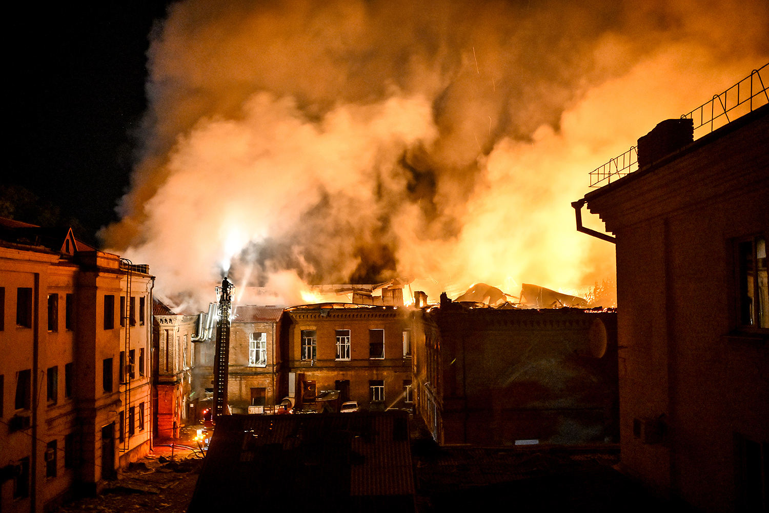 Flames and dense smoke engulf rooftops as firefighters battle a nighttime blaze among urban buildings.