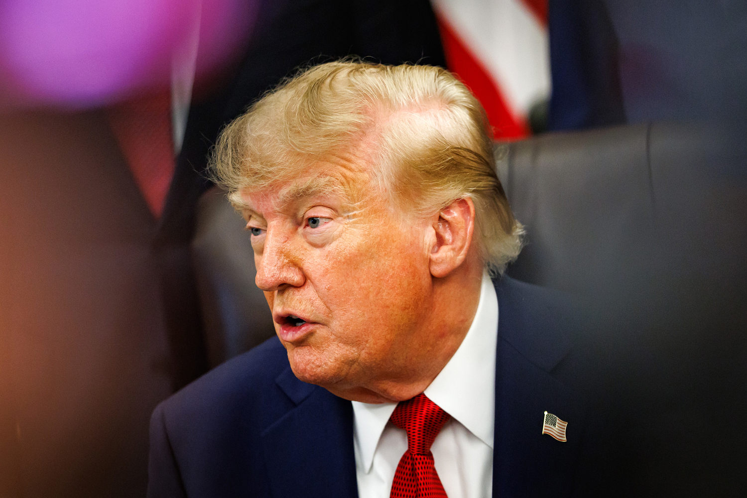 Trump, wearing a dark suit and red tie, speaks while seated with an American flag behind him.