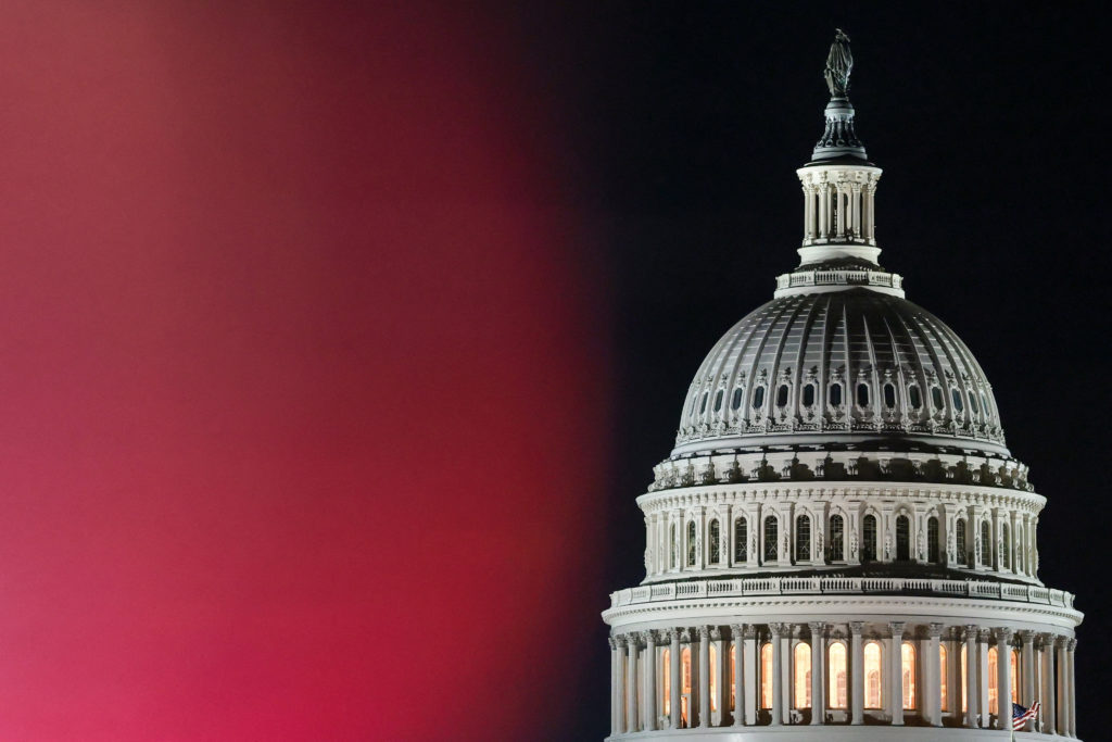 The U.S. Capitol dome is lit at night against a black sky, beside a blurred band of red light.