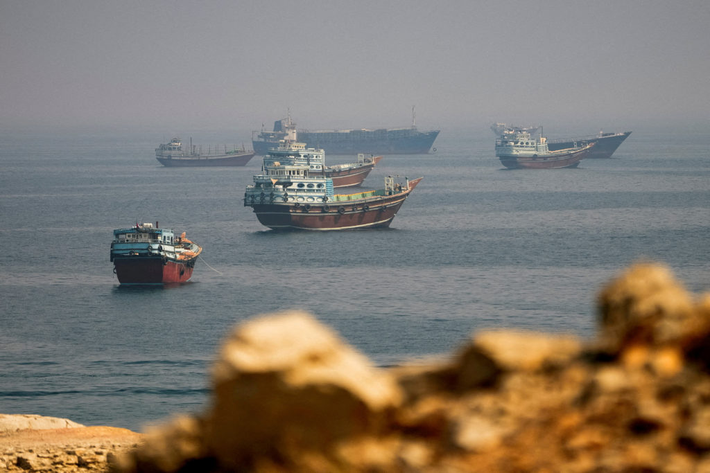 Several cargo vessels and wooden dhows sit at anchor on hazy water beyond a rocky shoreline.