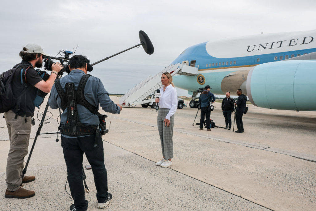 A television crew records a reporter standing on an airport tarmac beside the presidential aircraft and its boarding stairs.