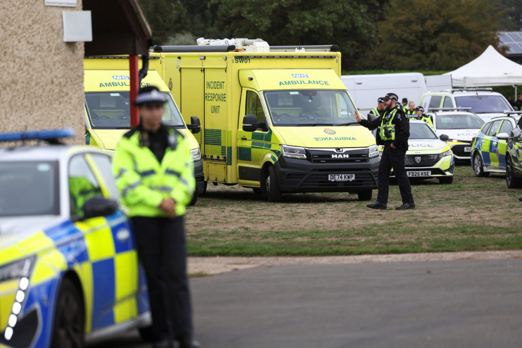 Police officers in high-visibility jackets stand beside ambulances, an incident response unit and patrol cars parked on grass.