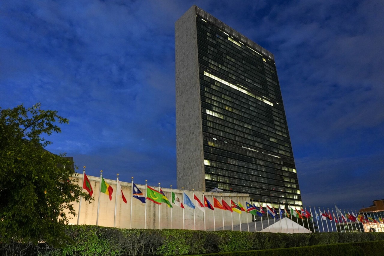 The United Nations Secretariat tower rises at dusk above a long row of national flags.