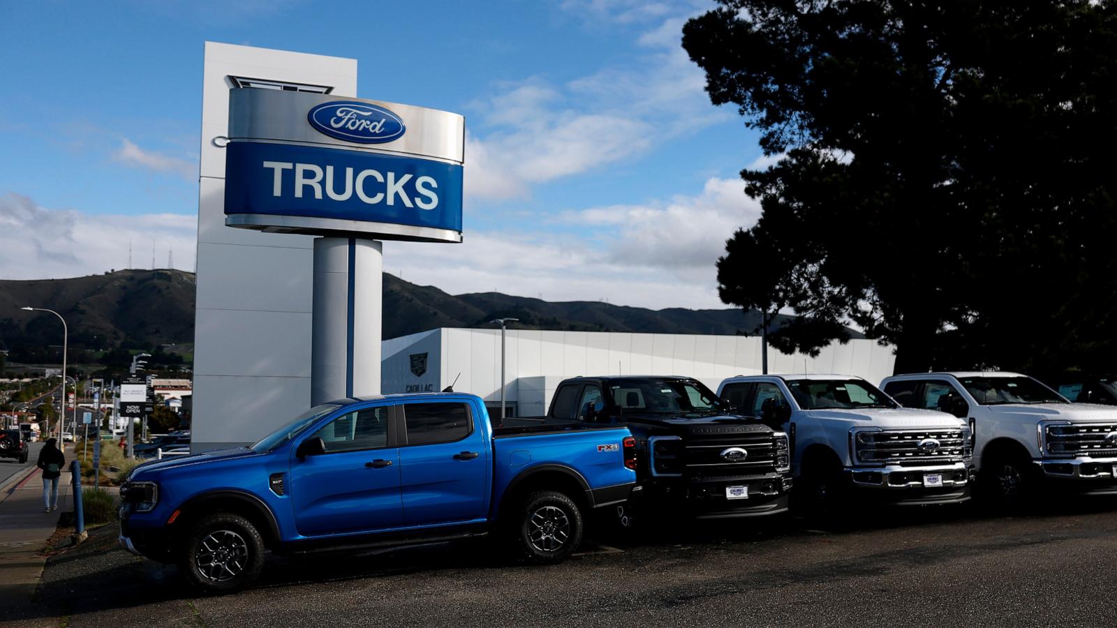 Pickup trucks stand in a row on a dealership lot beneath a tall Ford Trucks sign.