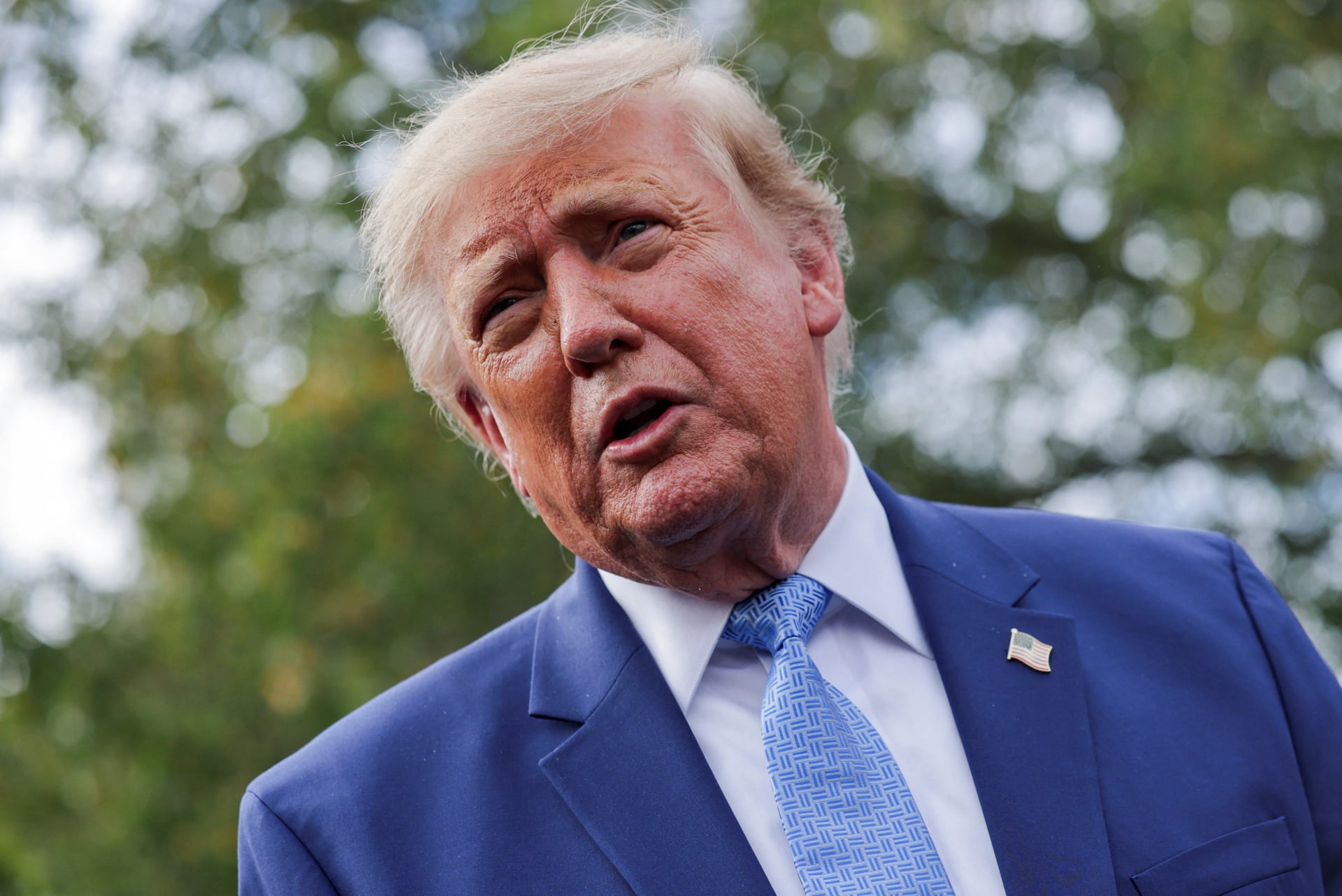 Trump, in a blue suit and patterned tie, speaks outdoors with trees blurred behind him.