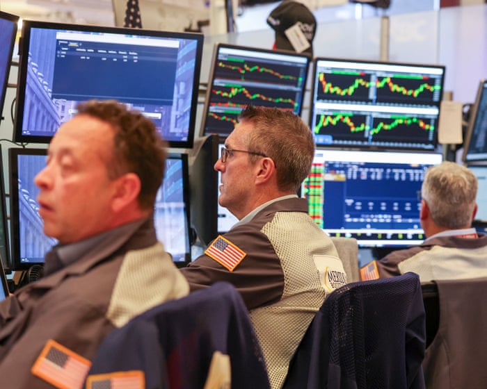 Traders in floor jackets sit before banks of monitors showing market charts.