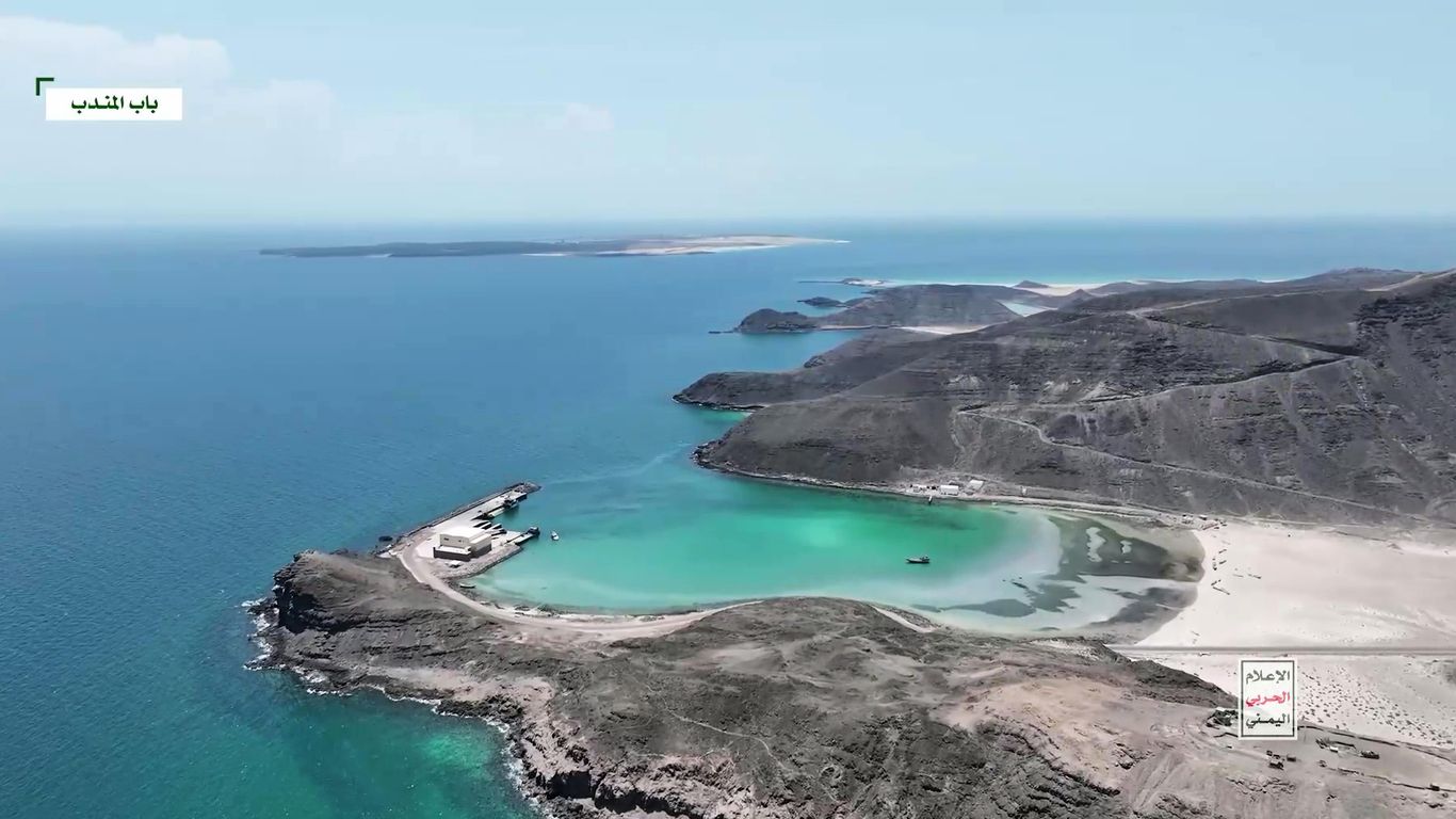 An aerial view of a rocky coastline with a turquoise bay, a pier and a sandy beach, islands on the horizon, with Arabic on-screen labels.
