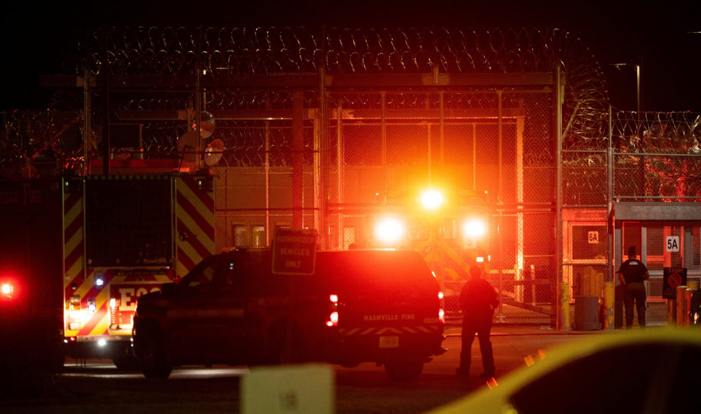 At night, a fire truck and an emergency vehicle with flashing lights wait inside a razor-wire prison fence as an officer stands by the gate.