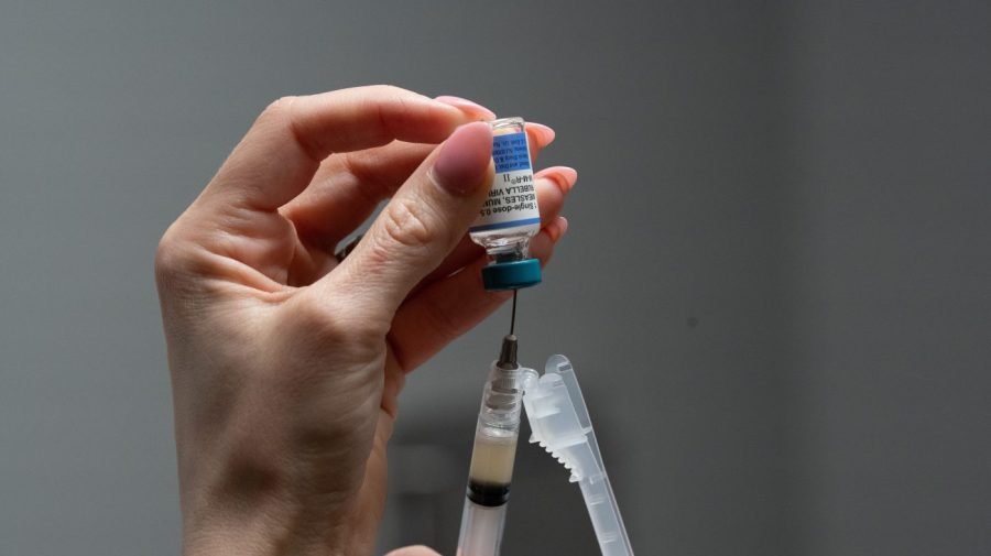 A hand holds a vaccine vial upside down while drawing liquid into a syringe.