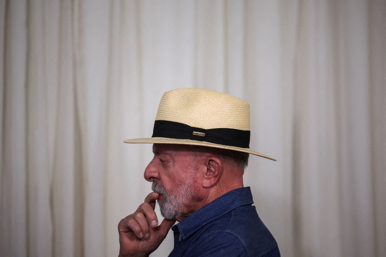 Lula, wearing a straw hat and blue shirt, rests a finger near his mouth against a plain curtain backdrop.