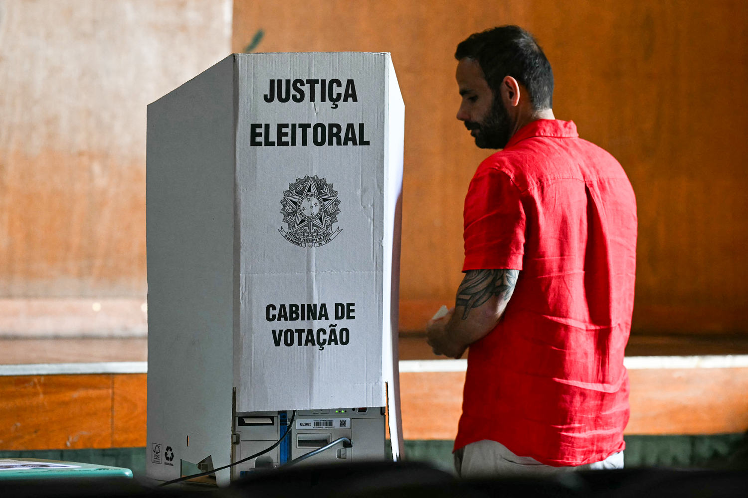 A man in a red shirt stands at a cardboard voting booth marked Justiça Eleitoral, Cabina de Votação.