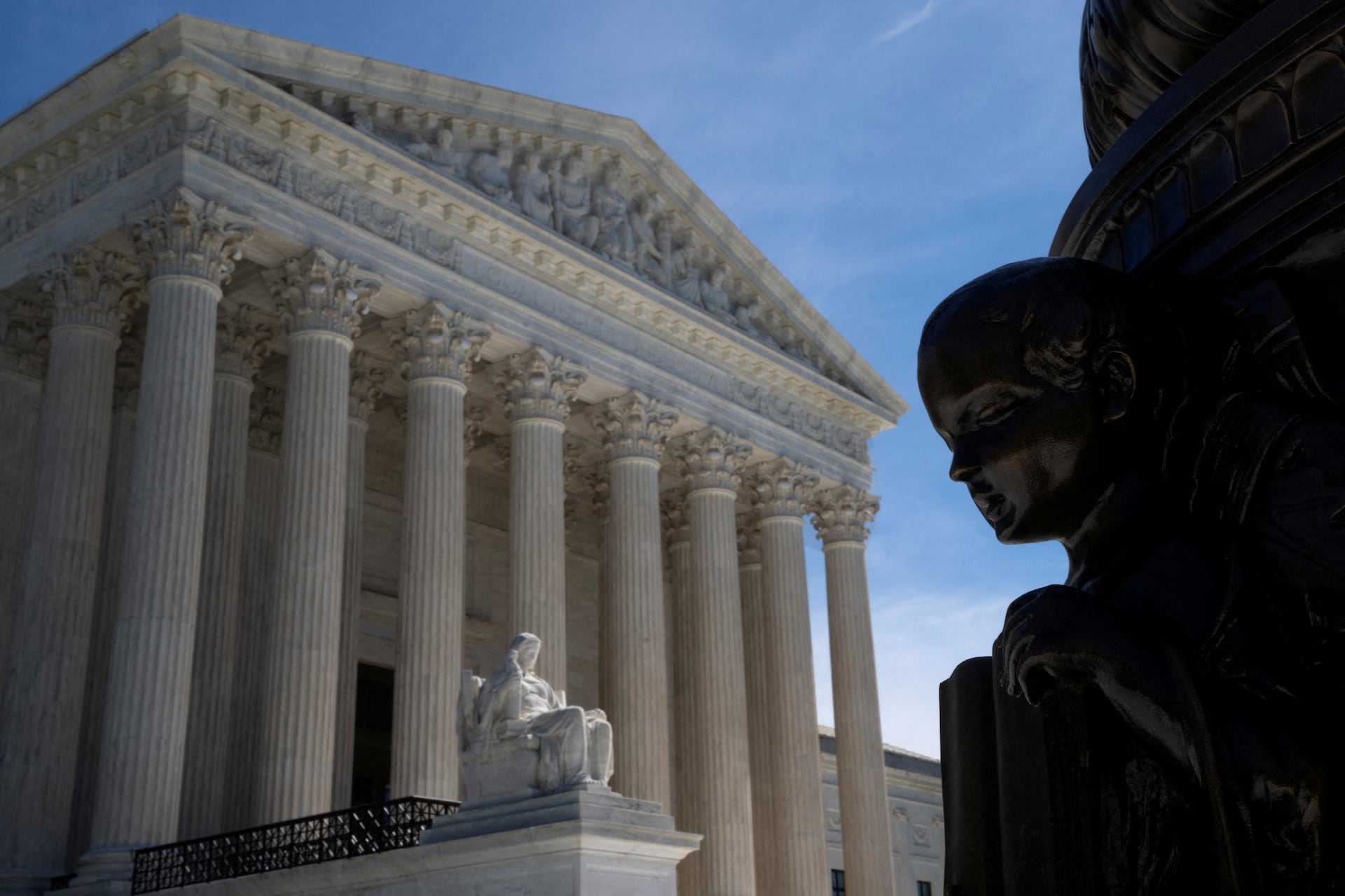 The Supreme Court's columned facade in sunlight under a deep blue sky, beside a statue in shadow.