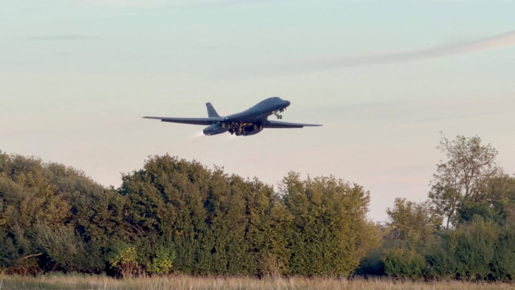 A military aircraft with landing gear extended flies low above a line of trees.