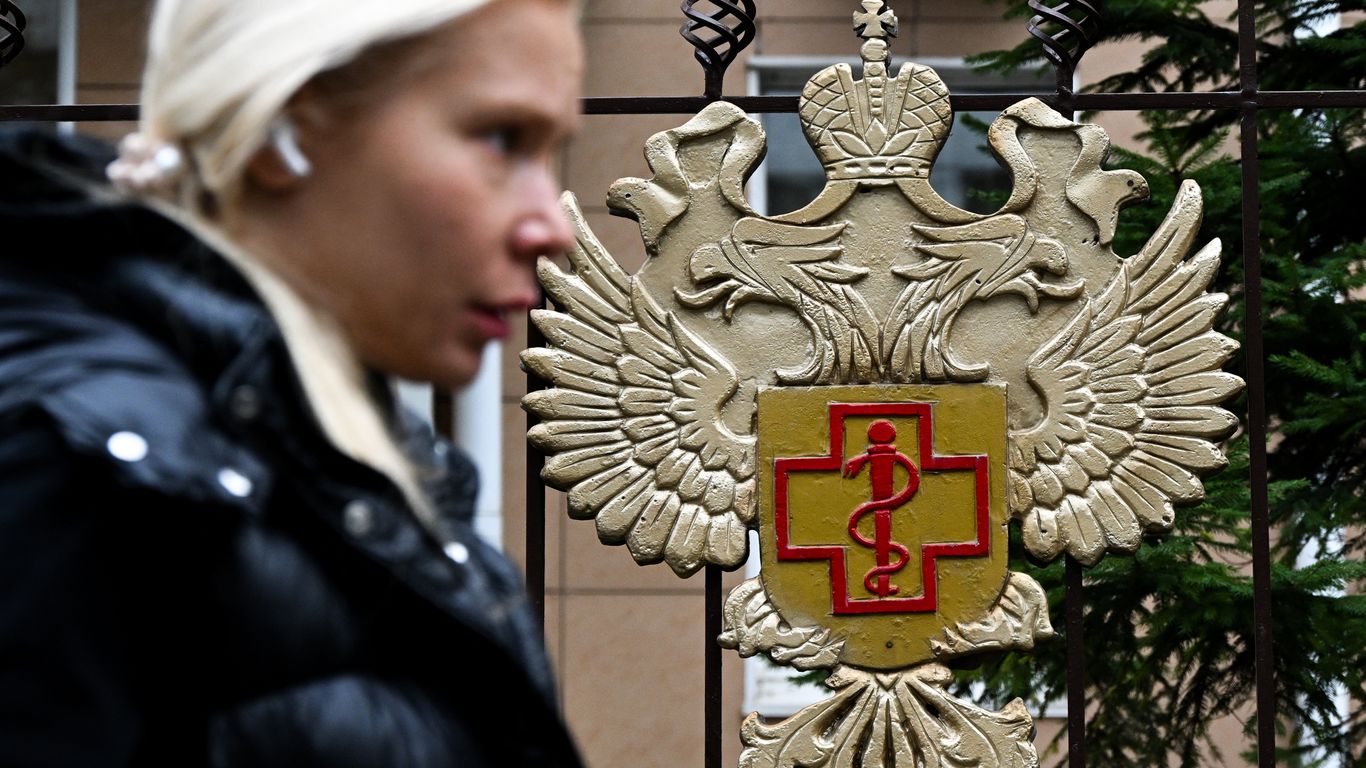 A blurred woman walks past a metal Russian double-headed eagle emblem bearing a red medical cross.
