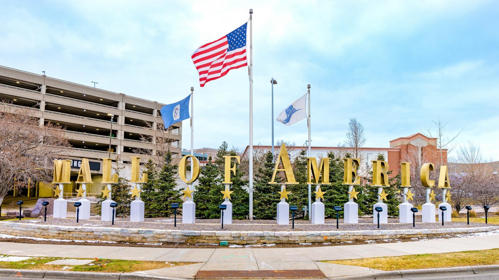 Gold letters spelling Mall of America stand on white pedestals in front of evergreen trees, beneath a U.S. flag and two other flags, beside a parking garage.
