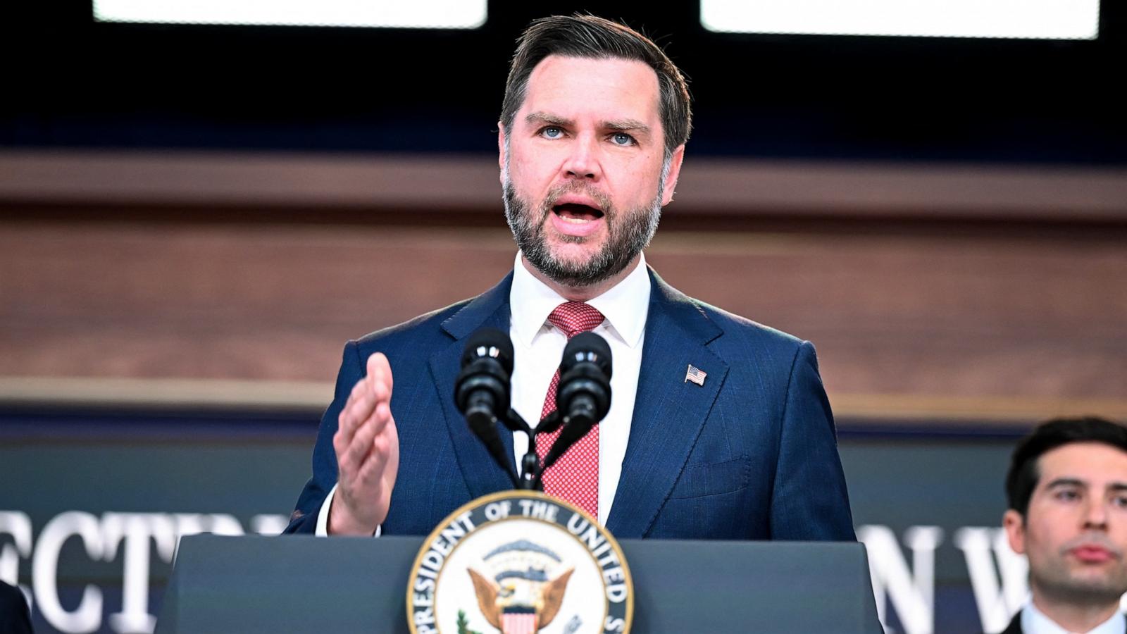 JD Vance, in a blue suit and red tie, speaks at a lectern bearing the vice-presidential seal and raises one hand, with a young man in a suit behind him at right.