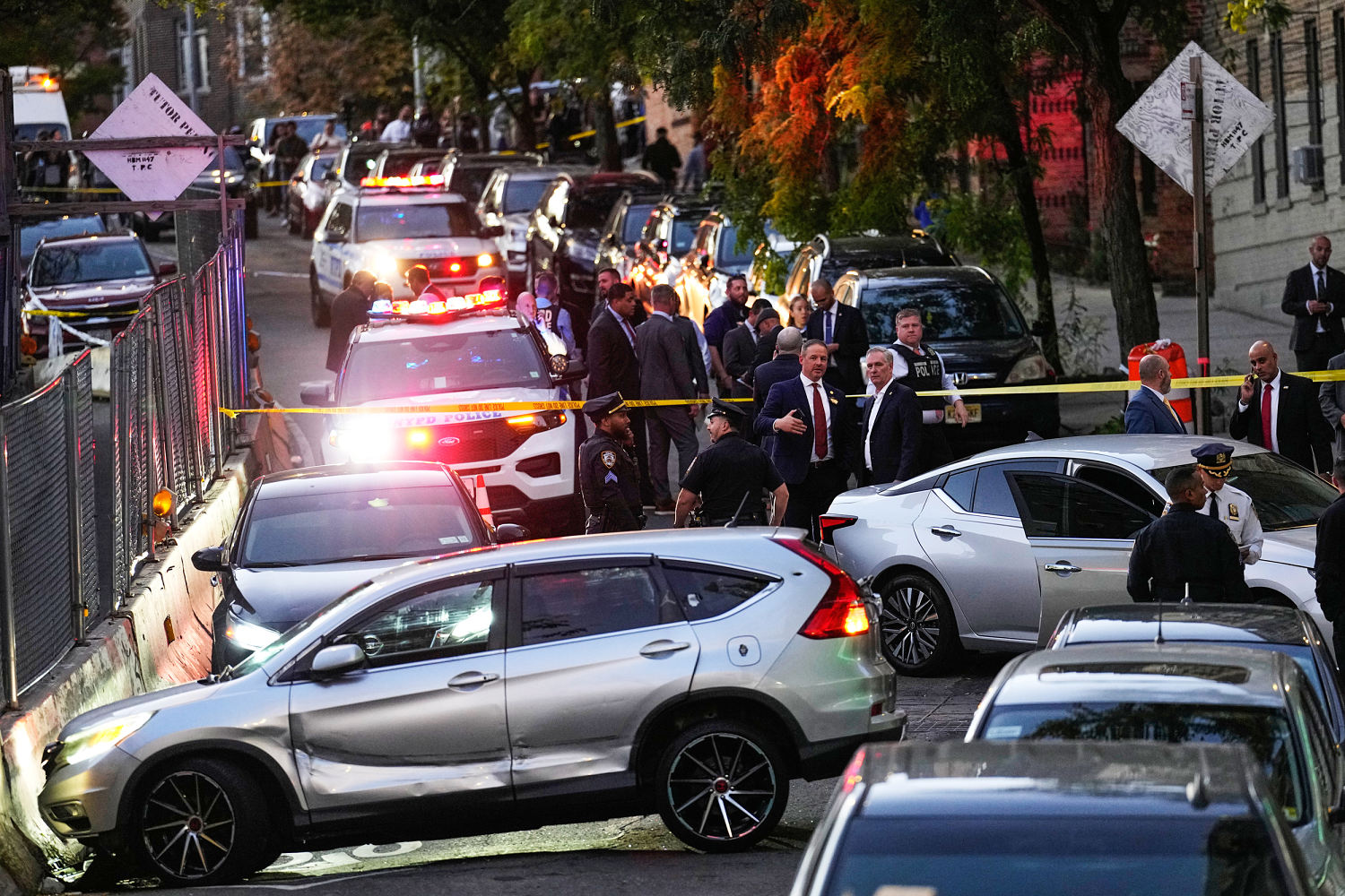 At dusk on a narrow residential street, NYPD vehicles with flashing lights stand behind yellow police tape, with uniformed officers and men in suits gathered around a silver SUV and a silver sedan.