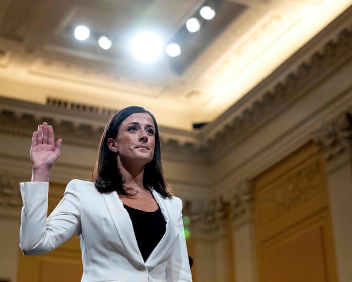Cassidy Hutchinson, in a white blazer over a black top, raises her right hand beneath bright ceiling lights in an ornate hearing room.