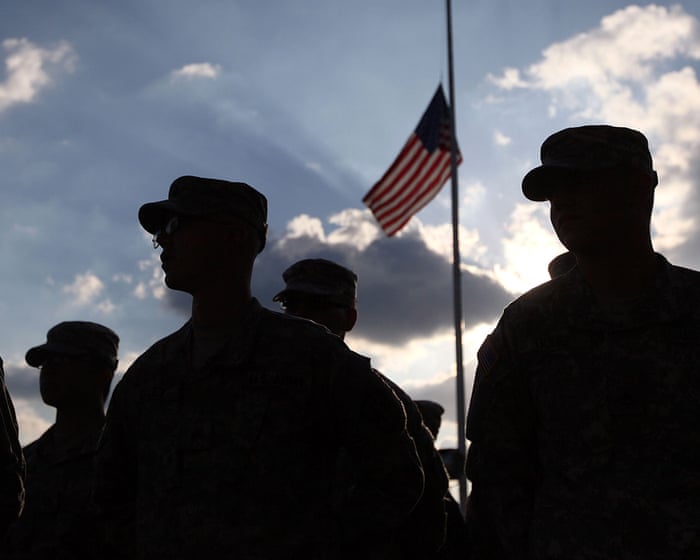 Soldiers in patrol caps stand in silhouette beneath an American flag flying at half-staff against a bright, cloudy sky.
