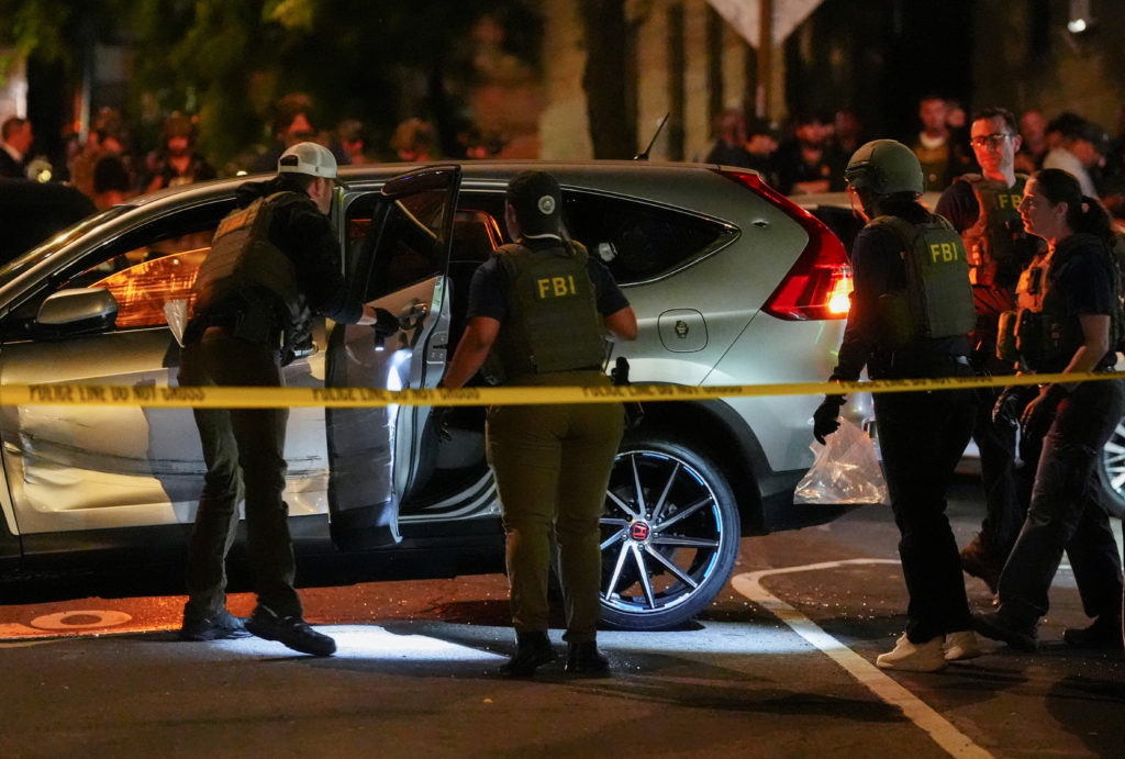 At night, behind yellow police tape, investigators in FBI vests stand around a silver SUV with its driver's door open and a dent along its side.