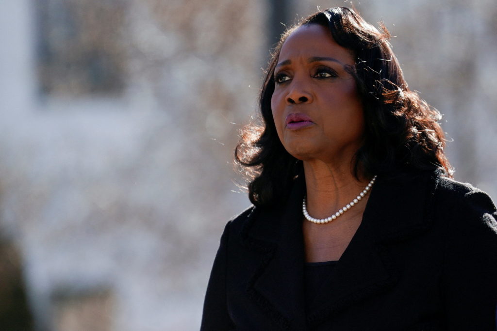 Lisa Cook, in a black coat and a pearl necklace, stands outdoors in bright winter sun, with a blurred stone building behind her.