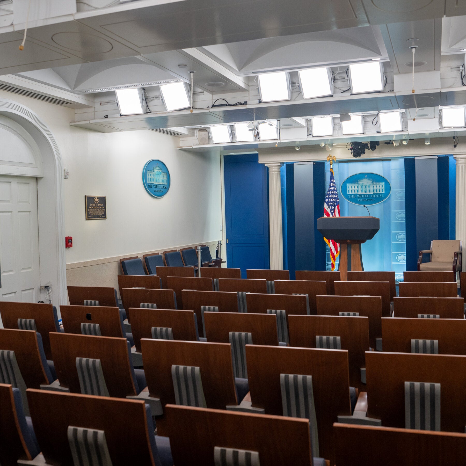The empty White House briefing room: rows of brown seats face a lectern, an American flag and the White House seal beneath a ceiling of television lights.