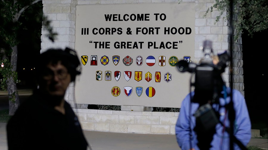 At night, a television camera on a tripod and a crew member stand before the lit Fort Hood welcome sign, which reads “Welcome to III Corps & Fort Hood, ‘The Great Place.’”