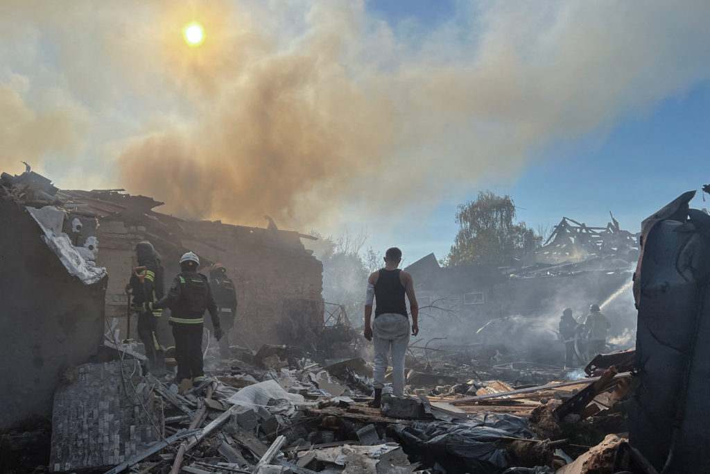 Firefighters work among smoking rubble and wrecked buildings under a sky thick with smoke, while a man in a black sleeveless shirt stands in the debris.