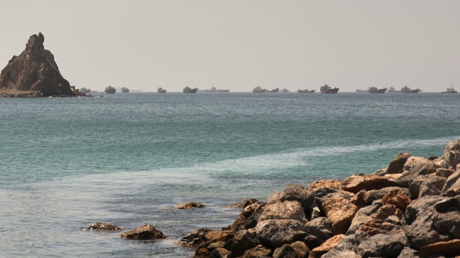 A long line of cargo ships rides at anchor on hazy water beyond a rocky shore, with a tall rock outcrop at the left.