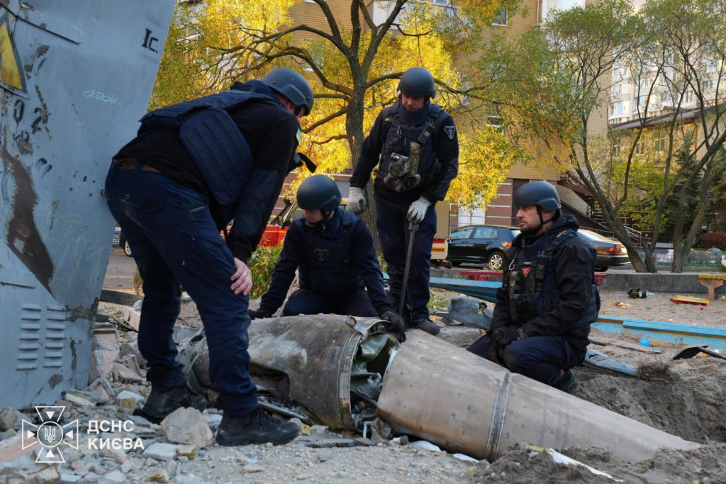 Four helmeted emergency workers gather around large cylindrical metal wreckage amid rubble, with trees and residential buildings behind them.