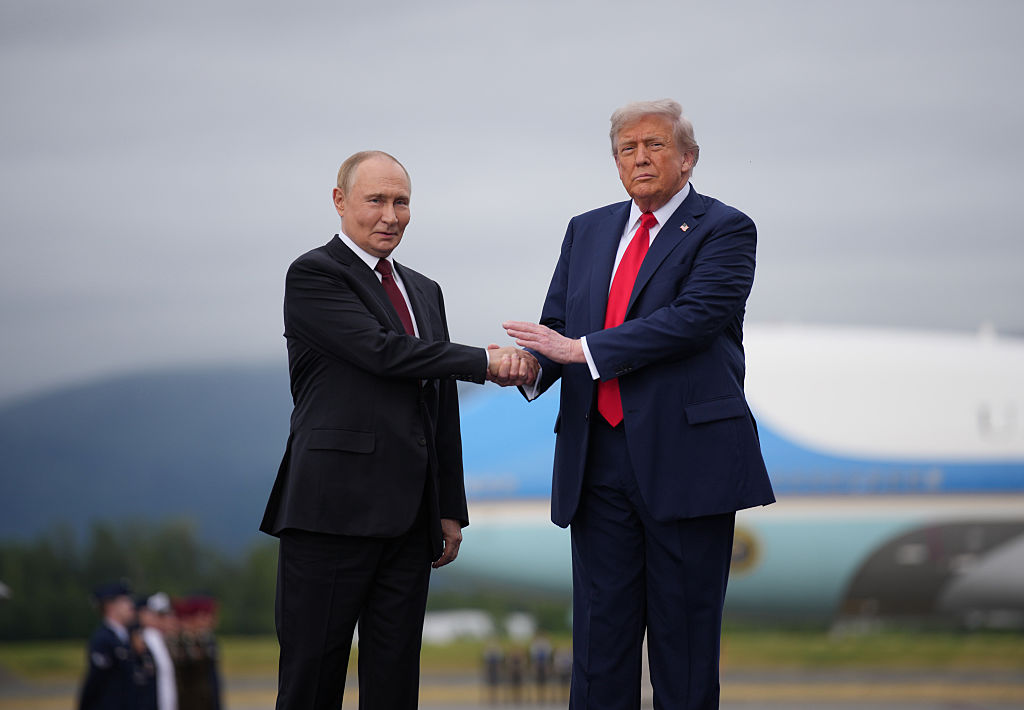 Vladimir Putin and Donald Trump, in dark suits, shake hands on an airfield under a gray sky, with a large aircraft behind them.