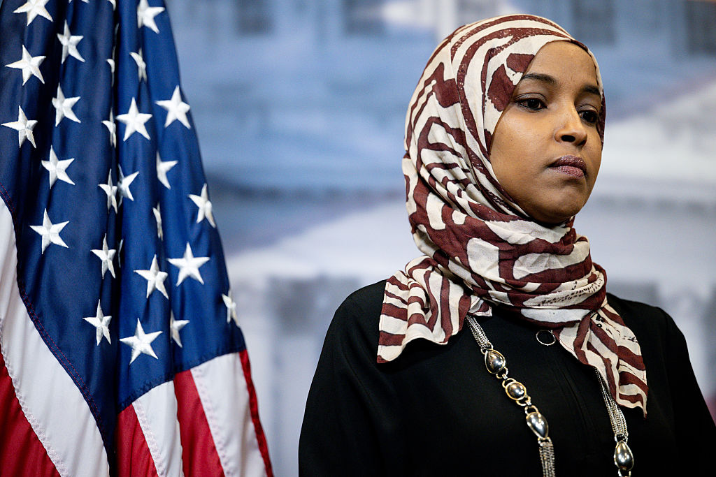 Ilhan Omar, in a burgundy-and-white patterned headscarf and black clothing, stands beside an American flag.